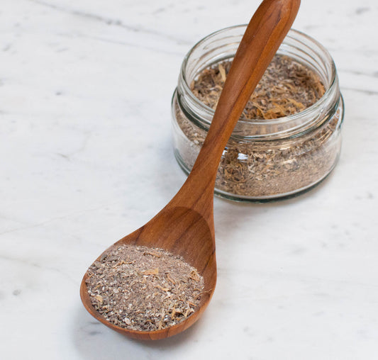 A wooden spoonful of rhassoul clay cleanser, with more of the product visible in the background behind the jar.